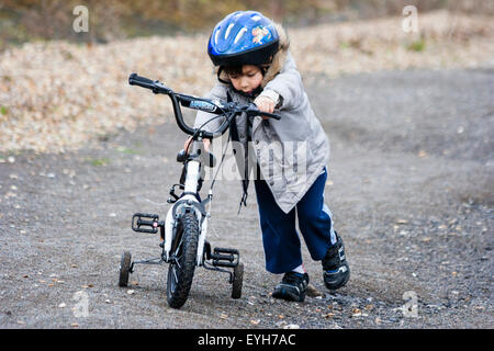 Boy struggling to push bicycle with stabilizers, up rough track in the winter. Caucasian child, 6-7 year old, with coat and crash helmet. Stock Photo