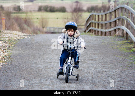 Boy struggling to ride bicycle with stabilizers, on rough track in the winter. Caucasian child, 6-7 year old, with coat and crash helmet. Wood fence. Stock Photo