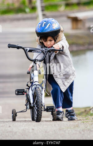 Boy struggling to push bicycle with stabilizers, up rough track in the winter. Caucasian child, 6-7 year old, with coat and crash helmet. Stock Photo