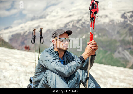 Young man mountaineer sitting on a snow hillside near top of mount Cheget and holding trekking pole. Mountain Elbrus on backgrou Stock Photo