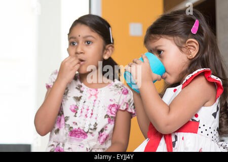 Two cute Indian girls eating food and drinking water. Asian sibling or children enjoying tea time food, living lifestyle at home Stock Photo