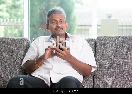 Smartphone mobile apps concept. Indian mature guy using mobile phone. Asian man relaxed and sitting on sofa indoor. Stock Photo