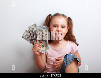 Funny small kid girl holding money and showing the tongue on blue background Stock Photo