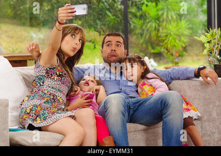 Family portrait of father, mother and two daughters sitting together in sofa posing for selfie making funny faces Stock Photo