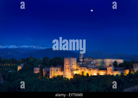 Moonrise over lit hilltop Alhambra Palace fortress complex at twilight Granada with snow capped Sierra Nevada Mountains Spain Stock Photo
