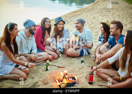 Young friends sitting around campfire and singing by guitar in the evening Stock Photo