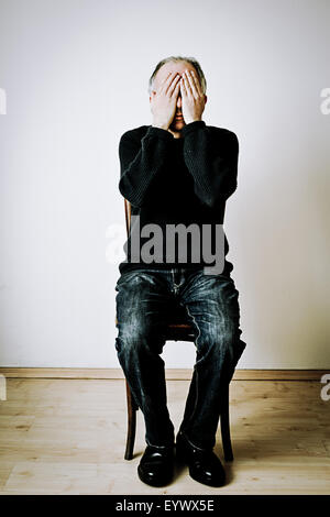 man sitting on a chair in a room with hands covering his face Stock Photo