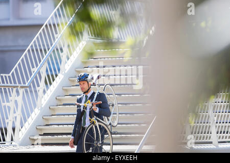 Businessman in suit and helmet carrying bicycle down urban stairs Stock Photo