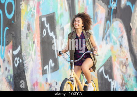 Smiling brunette woman riding bicycle along urban multicolor graffiti wall Stock Photo
