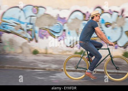 Hipster man riding bicycle on road along urban graffiti wall Stock Photo