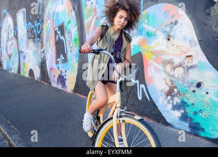 Portrait serious woman with afro on bicycle next to urban graffiti wall
