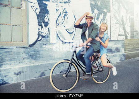 Portrait playful couple riding bicycle along urban graffiti wall Stock Photo