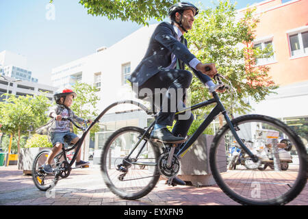 Businessman father in suit riding tandem bicycle with son on urban sidewalk Stock Photo