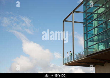 Men talking on balcony of glass bump out against blue sky Stock Photo