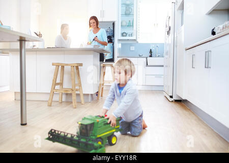 Women cooking in kitchen while boy plays with toy tractor on floor Stock Photo