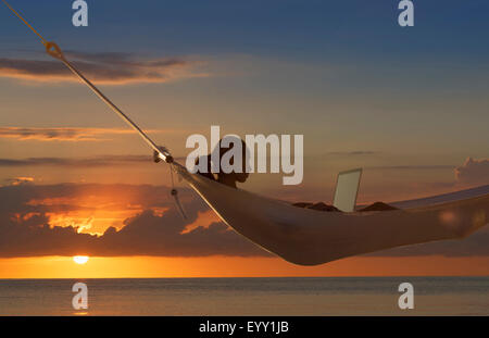Caucasian woman using laptop in hammock near ocean at sunset Stock Photo