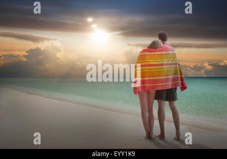 Caucasian couple wrapped in towel admiring sunset on beach Stock Photo