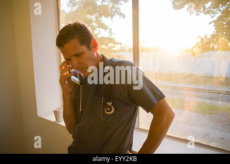 Hispanic doctor talking on cell phone at window Stock Photo