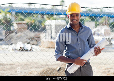 Black architect in hard hat holding blueprints near fence Stock Photo