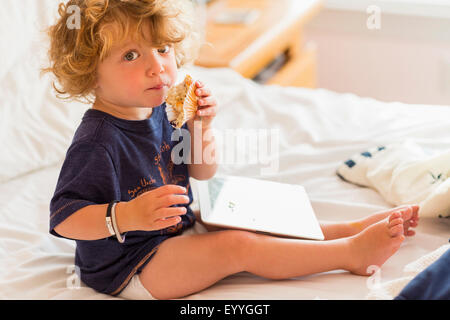 Caucasian baby boy eating and using digital tablet on bed Stock Photo