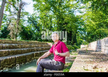 Charismatic and handsome Caucasian sportsman of forty with grey hair wearing red polo shirt and dark linen trousers indicates himself sitting on the ancient medieval steps of Mill Canal in Emilia Romagna in Italy Stock Photo