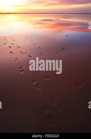 footprints on beach at outgoing tide and reflection of clouds, mudflats at sunset, United Kingdom Stock Photo