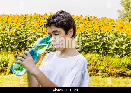Environmentally-friendly living - Caucasian boy in a field of sunflowers is about to drink clear water from a plastic bottle green bluish to quench thirst on a warm and bright summer day Stock Photo