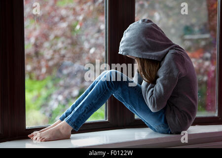 Sad girl in hooded top, sitting on a window ledge with head in hands in despair Stock Photo