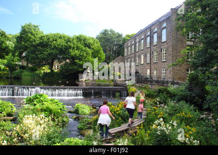 People walk over the wooden public footbridge by Bamford Mill and weir in the Peak District Derbyshire England UK - summer Stock Photo