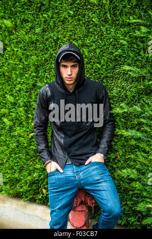 Handsome young man in black hoodie sweater standing outdoor against green plant hedge looking at camera Stock Photo