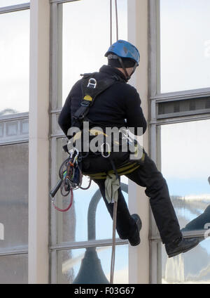 High rise window cleaner hangs from rope wearing harness on outside of office building Stock Photo