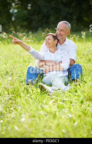 Senior couple sitting in grass in nature and woman pointing into the distance Stock Photo