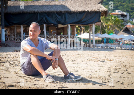 Tourist in Puerto Vallarta, Mexico. Man, 55 years old, hispanic ethnicity, sitting in the sand. Palapa restaurant in the backgro Stock Photo