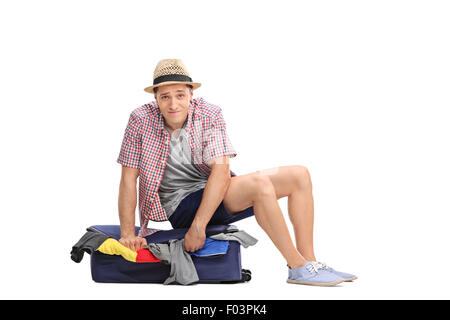 Sad young male tourist sitting on top of a packed suitcase and trying to close it isolated on white background Stock Photo