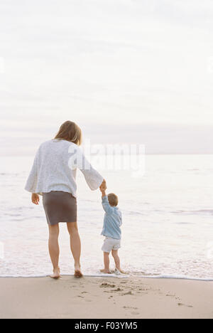 Woman standing on a sandy beach by the ocean, holding her young son's hand. Stock Photo