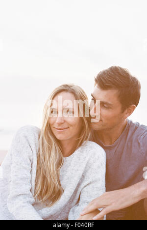 A couple sitting close on a beach, a man and woman with their arms around each other and heads together. Stock Photo