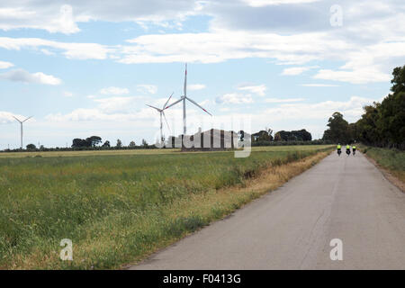 Three touring cyclists riding their bikes on a country road by a field of wheat and wind turbines. Stock Photo