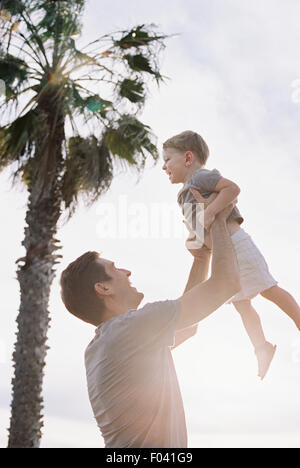 Man standing by a palm tree, playing with his young son, lifting him into the air. Stock Photo