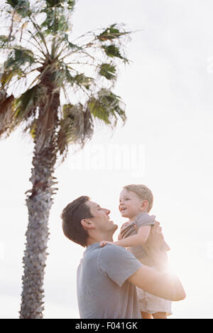 Smiling man standing by a palm tree, carrying his young son in his arms. Stock Photo