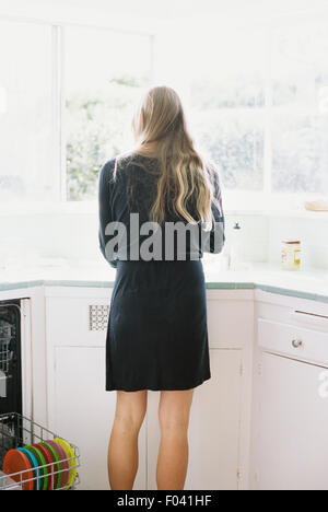 Rear view of a woman with long blond hair standing at a kitchen sink. Stock Photo