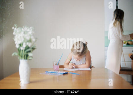 Young girl sitting at  a table, painting, a vase with white flowers, a woman in the background. Stock Photo