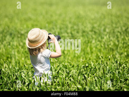 Little blonde girl in straw hat looking through binoculars outdoor