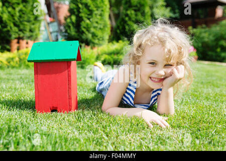 Wooden model of house and little blonde girl lying on green grass. Real estate concept Stock Photo