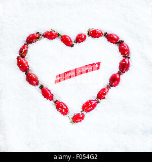 a heart-shaped wreath made with rose hips laid out on the snow, and piece of red ribbon saying Merry Christmas Stock Photo
