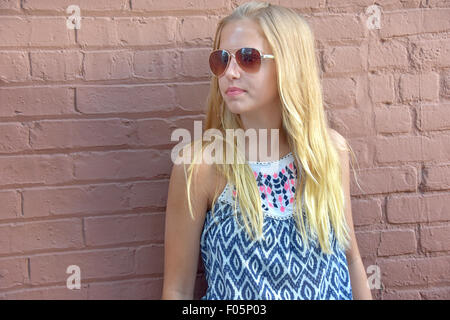 Young teenage girl wearing aviator sunglasses and leaning on painted brick wall. Stock Photo