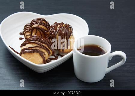 Close up Delicious Pastries with Chocolate Syrup on a White Heart Shaped Plate and a Cup of Tea on Top of a Gray Wooden Table Stock Photo