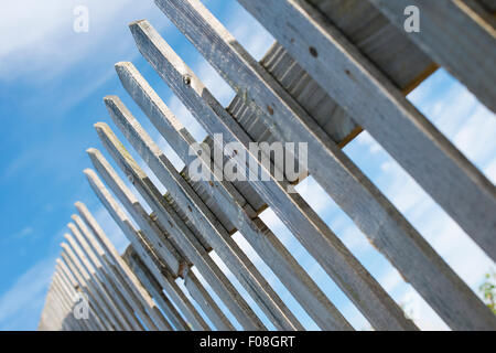 Low angle, tilted view of an old wooden picket fence, against a blue sky with clouds. Stock Photo