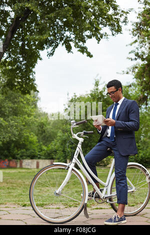 Young businessman sitting on bicycle and networking in park Stock Photo