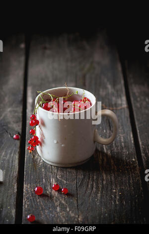 Old white cup full of fresh ripe red currant over old wooden table. Dark rustic style. Natural day light. Stock Photo