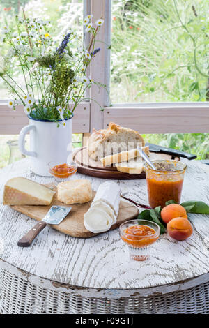 Cheese platter, rustic bread and chutney on white wooden table with flower bouquet and wooden boards Stock Photo